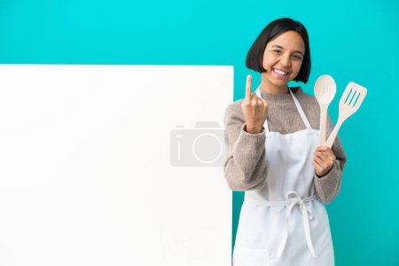Téléchargez les photos : Jeune femme cuisinière mixte avec une grande pancarte isolée sur fond bleu faisant geste à venir - en image libre de droit