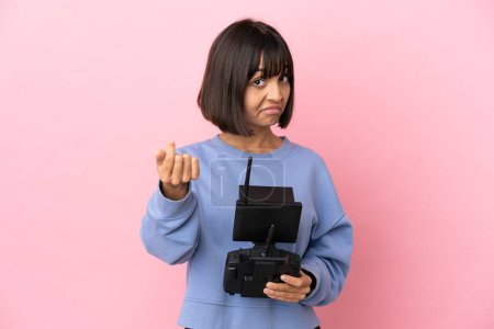 Joven mujer de raza mixta sosteniendo un control remoto de dron aislado sobre fondo rosa haciendo gesto de dinero