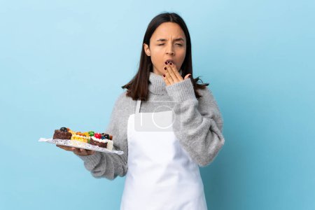 Pastry chef holding a big cake over isolated blue background yawning and covering wide open mouth with hand.