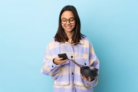 Téléchargez les photos : Jeune femme brune métisse tenant un bol rempli de nouilles sur fond bleu isolé envoyant un message avec le mobile. - en image libre de droit