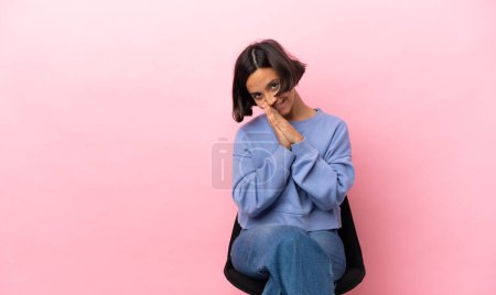 Mujer joven de raza mixta sentada en una silla aislada sobre fondo rosa mantiene unida la palma. Persona pide algo