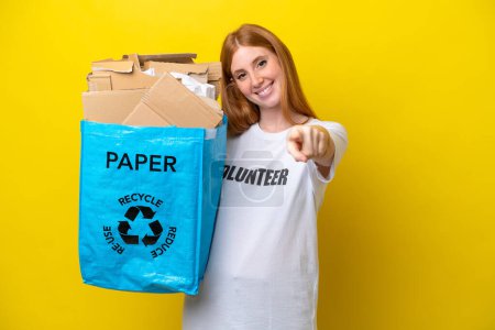 Foto de Joven pelirroja sosteniendo una bolsa de reciclaje llena de papel para reciclar aislada sobre fondo amarillo apuntando al frente con expresión feliz - Imagen libre de derechos