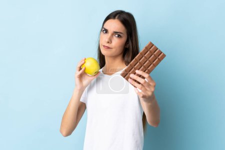 Photo for Young caucasian woman isolated on blue background taking a chocolate tablet in one hand and an apple in the other - Royalty Free Image