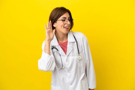 Foto de Mujer doctora aislada sobre fondo amarillo escuchando algo poniendo la mano en la oreja - Imagen libre de derechos