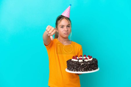 Téléchargez les photos : Petite fille caucasienne tenant gâteau d'anniversaire isolé sur fond bleu montrant pouce vers le bas avec expression négative - en image libre de droit