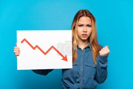 Foto de Teenager girl over isolated blue background holding a sign with a decreasing statistics arrow symbol and angry - Imagen libre de derechos