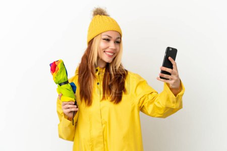 Blondes Mädchen in einem regendichten Mantel über isoliertem weißem Hintergrund macht ein Selfie