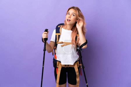 Téléchargez les photos : Adolescente avec sac à dos et bâtons de trekking sur fond violet isolé écoutant quelque chose en mettant la main sur l'oreille - en image libre de droit