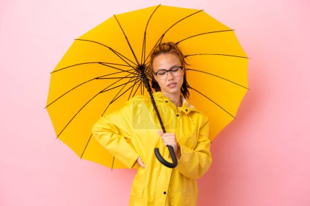 Teenager russische Mädchen mit regendichtem Mantel und Regenschirm isoliert auf rosa Hintergrund leiden unter Rückenschmerzen, weil sie sich bemüht haben