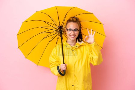 Teenager russische Mädchen mit regendichtem Mantel und Regenschirm isoliert auf rosa Hintergrund zeigt ok Zeichen mit den Fingern
