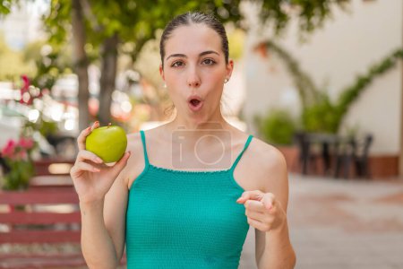 Téléchargez les photos : Jeune jolie femme avec une pomme à l'extérieur surpris et pointant devant - en image libre de droit
