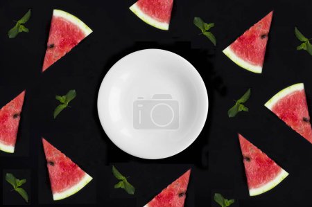 Téléchargez les photos : Assiette blanche vide pour texte et tranche de pastèque sur fond noir. Vue de dessus. Espace de copie. - en image libre de droit