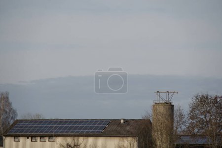 Téléchargez les photos : Solar modules on the roof of a large barn - electricity producer farm. The sun shines brightly on the roof and lets the modules shimmer in the light: the sky above is cloudy - en image libre de droit