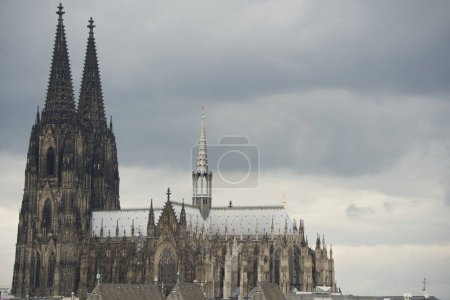 Der Kölner Dom in Seitenansicht. Der Dom erhebt sich hoch über den Schierfer Dächern der davor liegenden Gebäude. Die dunklen Regenwolken bringen buchstäblich den Kreuzungsturm und das Metalldach des Kirchenschiffs zum Leuchten