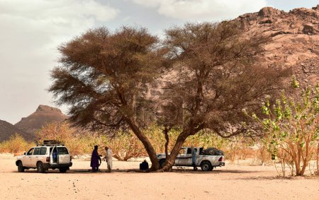 Foto de Djanet, Argelia - 2 de mayo de 2023: Un grupo de hombres tuareg con jeeps descansan bajo un árbol con el rocoso desierto del Sahara al fondo - Imagen libre de derechos