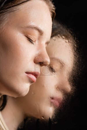 Foto de Vista de cerca de la mujer joven con la piel húmeda posando cerca del espejo sobre fondo negro - Imagen libre de derechos