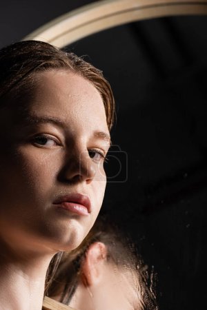 Junges Model mit nasser Haut im Gesicht blickt in Kamera in der Nähe von verschwommenem Spiegel 