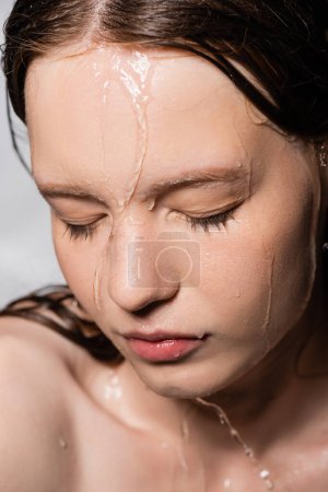 Close up view of water dripping on face of young woman isolated on grey 