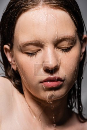 Vista de cerca del agua derramándose sobre la cara de la joven modelo aislada en gris 