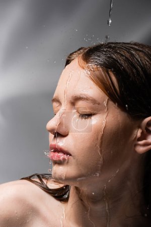 Young model with water on face and hair standing on abstract grey background 