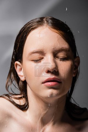 Junge Frau mit Wasser im Gesicht und Haaren, die die Augen vor abstraktem grauen Hintergrund schließen 