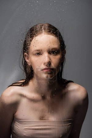 Young woman looking at camera behind wet glass on grey background 