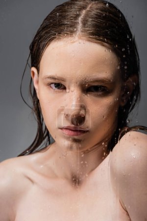 Portrait of woman with naked shoulders standing behind wet glass on grey background 