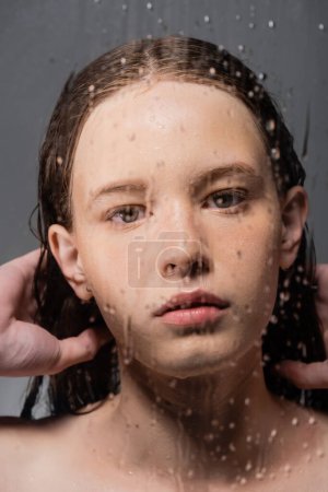 Junge Frau berührt Haare hinter nassem Glas auf grauem Hintergrund 