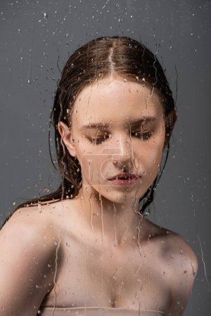 Foto de Modelo de pelo limpio con hombros desnudos detrás de vidrio mojado sobre fondo gris - Imagen libre de derechos