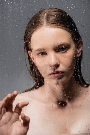 Young model with naked shoulders touching wet glass on grey background 