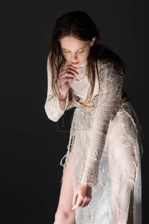 Stylish young model in fishnet skirt and knitted cardigan looking down isolated on black with light 
