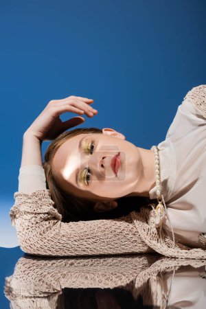 Foto de Sirena en punto cárdigan y collar de perlas mirando a la cámara cerca de la superficie reflectante sobre fondo azul degradado - Imagen libre de derechos