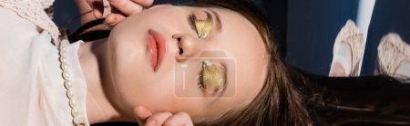 Top view of woman with golden makeup and pearl necklace lying on abstract background, banner 