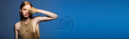 Téléchargez les photos : Jeune femme avec de la peinture dorée à la main couvrant l'oeil isolé sur bleu, bannière - en image libre de droit
