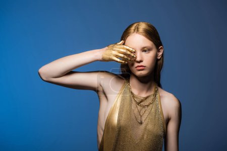 Mujer joven con pintura dorada en el ojo que cubre la mano aislado en azul 