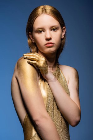 Porträt eines jungen Models in goldenem Kleid und Farbe, das isoliert auf blauem Grund in die Kamera blickt 
