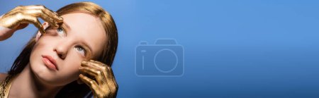 Téléchargez les photos : Portrait de jeune femme avec teinture dorée sur les mains et les cheveux touchant visage isolé sur bleu, bannière - en image libre de droit