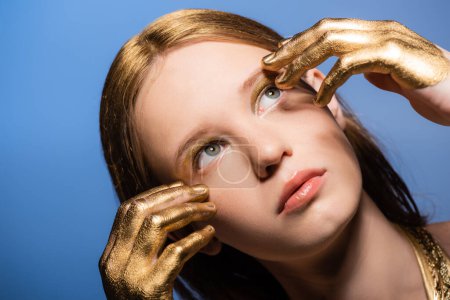 Téléchargez les photos : Jolie jeune femme avec de la peinture dorée sur les mains touchant le visage et regardant loin isolé sur le bleu - en image libre de droit