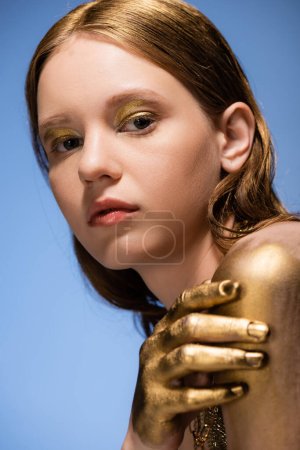 Portrait of young woman with golden makeup and paint on hand looking at camera isolated on blue 