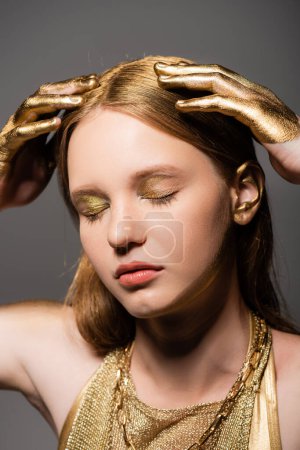 Modelo joven con maquillaje y manos en pintura dorada tocando el cabello aislado en gris 