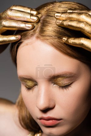 Portrait of young woman with golden makeup and paint on hands touching hair and closing eyes isolated on grey 