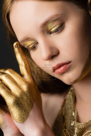 Portrait of young woman with golden makeup and paint on hand touching eyebrow isolated on grey 