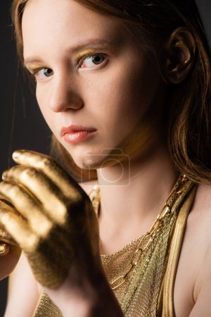Portrait of woman with golden paint on hand and face looking at camera isolated on grey 