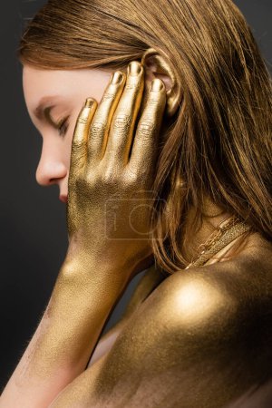 Foto de Vista lateral de mujer joven en pintura dorada tocando oreja aislada en gris - Imagen libre de derechos