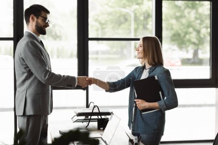 Photo for Side view of smiling businessman shaking hand of young woman with resume in office - Royalty Free Image
