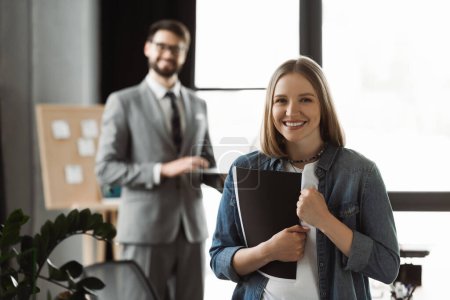 Cheerful woman with resume showing yes gesture near blurred businessman in office 
