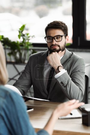 Businessman in eyeglasses holding resume and looking at blurred woman in office 