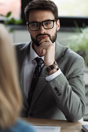 Hombre de negocios concentrado en traje mirando a la mujer borrosa durante la entrevista de trabajo en la oficina 