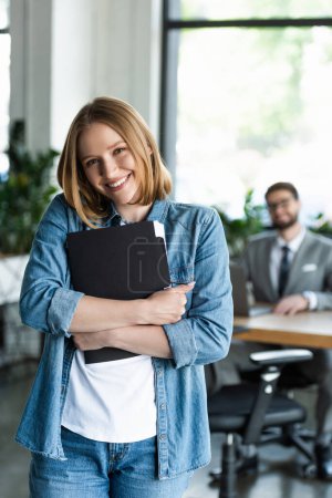 Glückliche junge Frau hält Papiermappe mit Lebenslauf in der Hand und blickt in die Kamera im Büro 