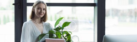 Positive intern looking at camera near plant and computer in office, banner 